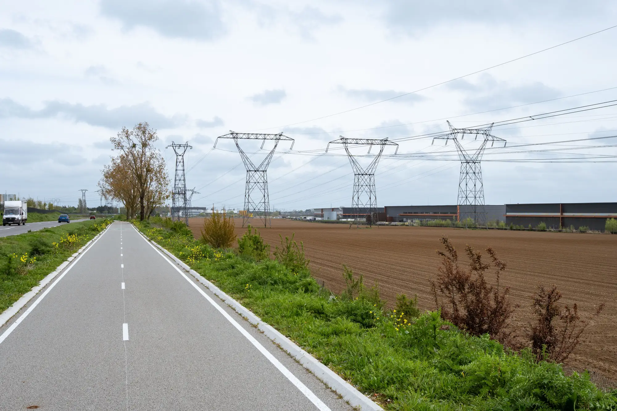 A ploughed field beside a road and a row of high-voltage pylons, with long warehouse buildings along its far edge